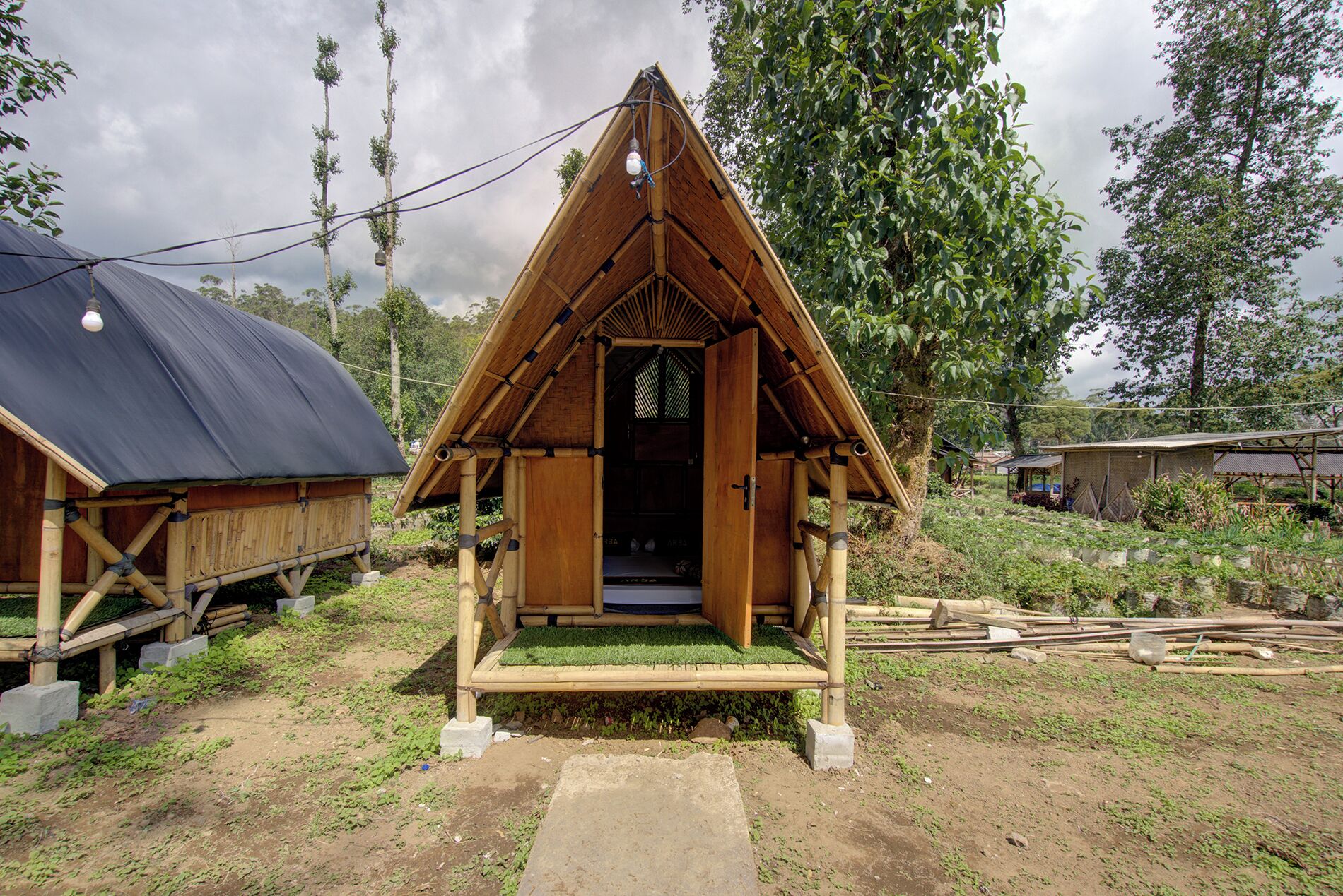 A small, wooden, triangular-roofed structure with a door, surrounded by greenery and a dirt path.