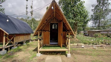 A small, wooden, triangular-roofed structure with a door, surrounded by greenery and a dirt path.