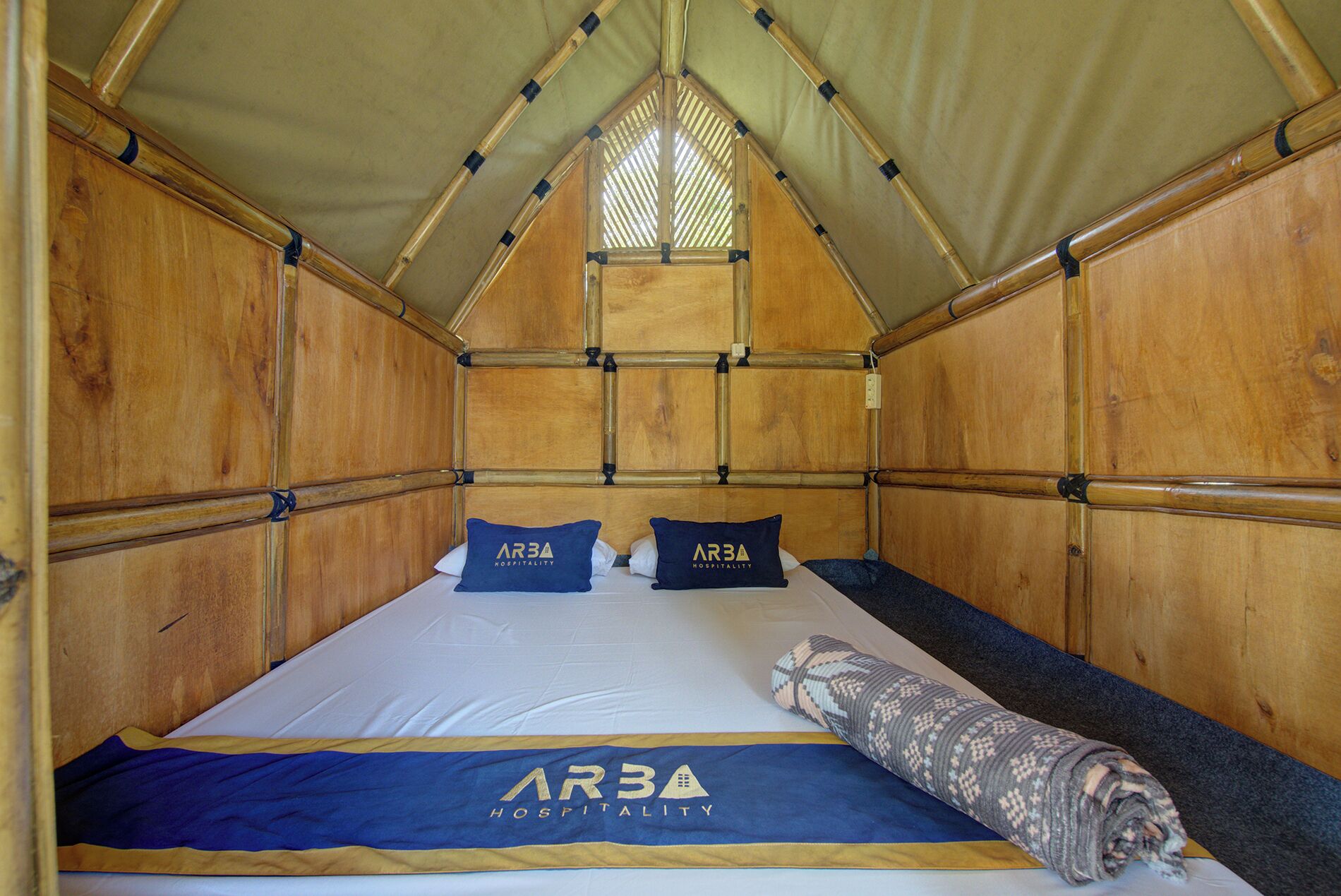 Un lit simple avec un baldaquin, deux oreillers portant la marque « ARBA HOSPITALITY » et une couverture à motifs.
