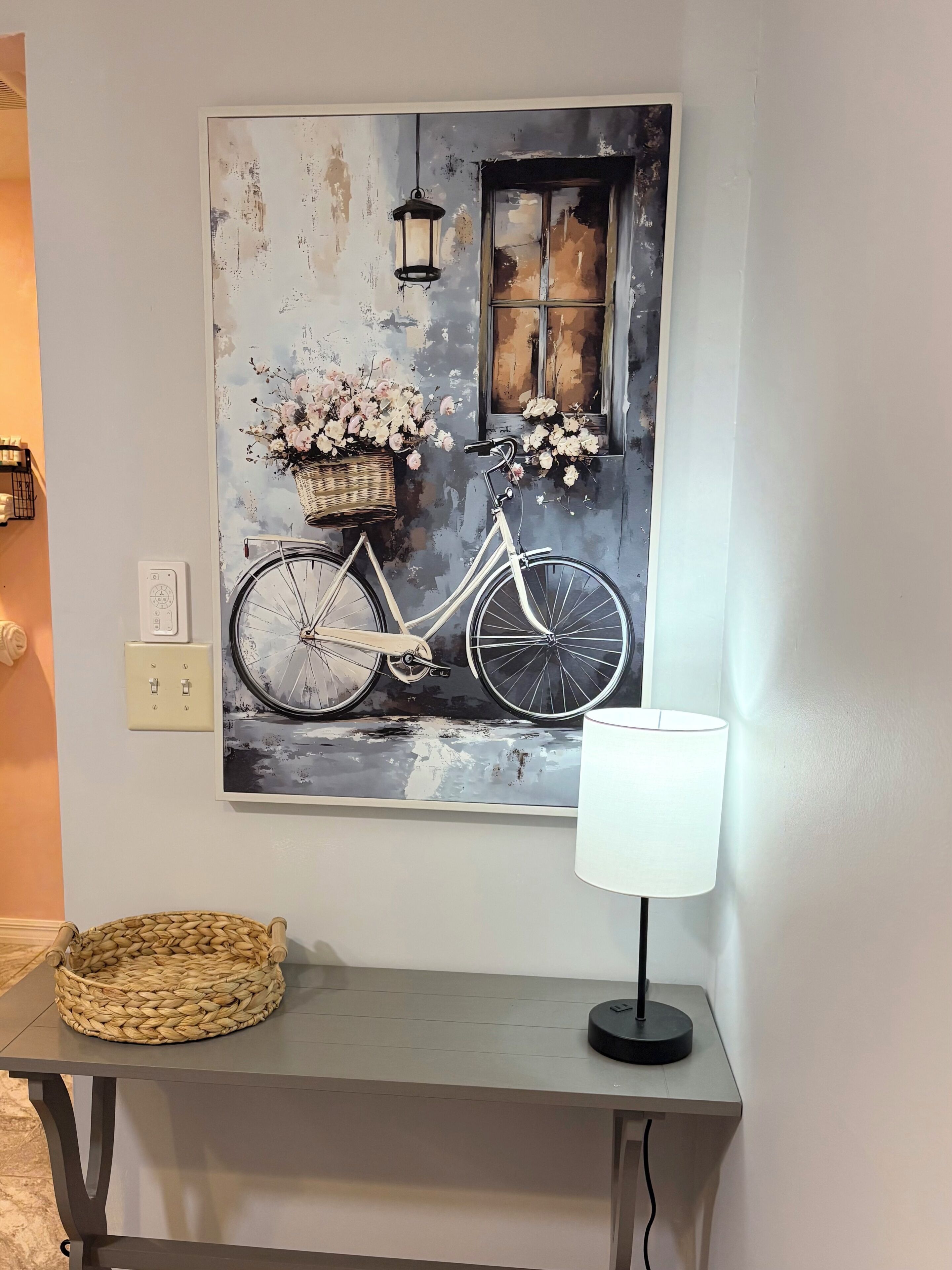 A wall-mounted painting of a bicycle with flowers, a basket, and a window, next to a table with a lamp.