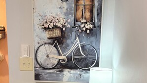 Un cuadro colgado en la pared muestra una bicicleta con flores, una cesta y una ventana, junto a una mesa con una lámpara.