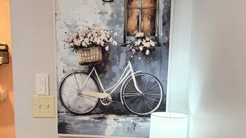 A wall-mounted painting of a bicycle with flowers, a basket, and a window, next to a table with a lamp.