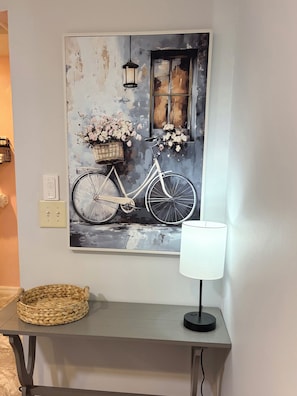Ein Wandbild zeigt ein Fahrrad mit Blumen, einem Korb und einem Fenster, neben einem Tisch mit einer Lampe.
