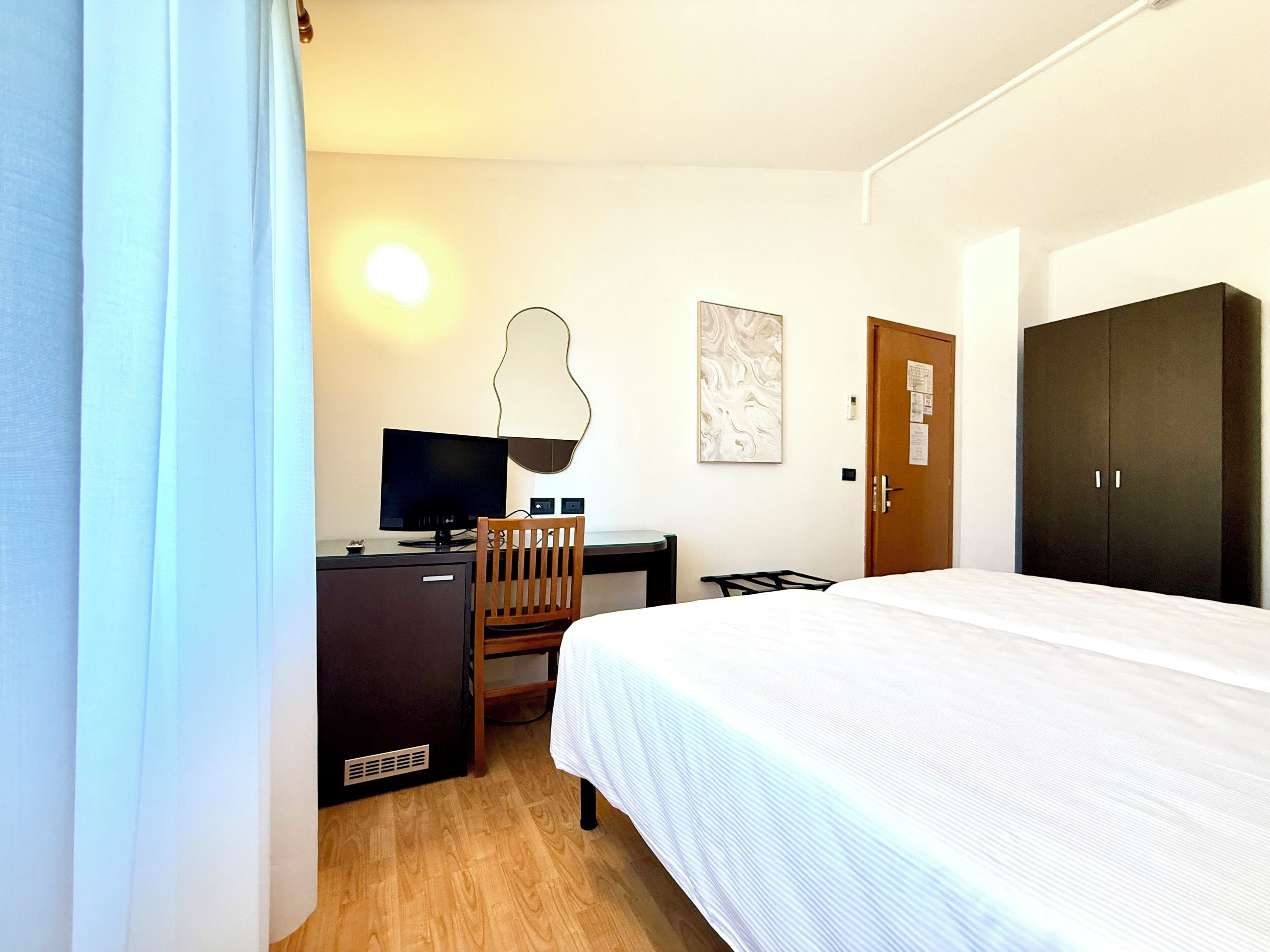 Una camera d'albergo con un letto, una scrivania con un computer, una sedia, un armadio e un'opera d'arte appesa al muro.
