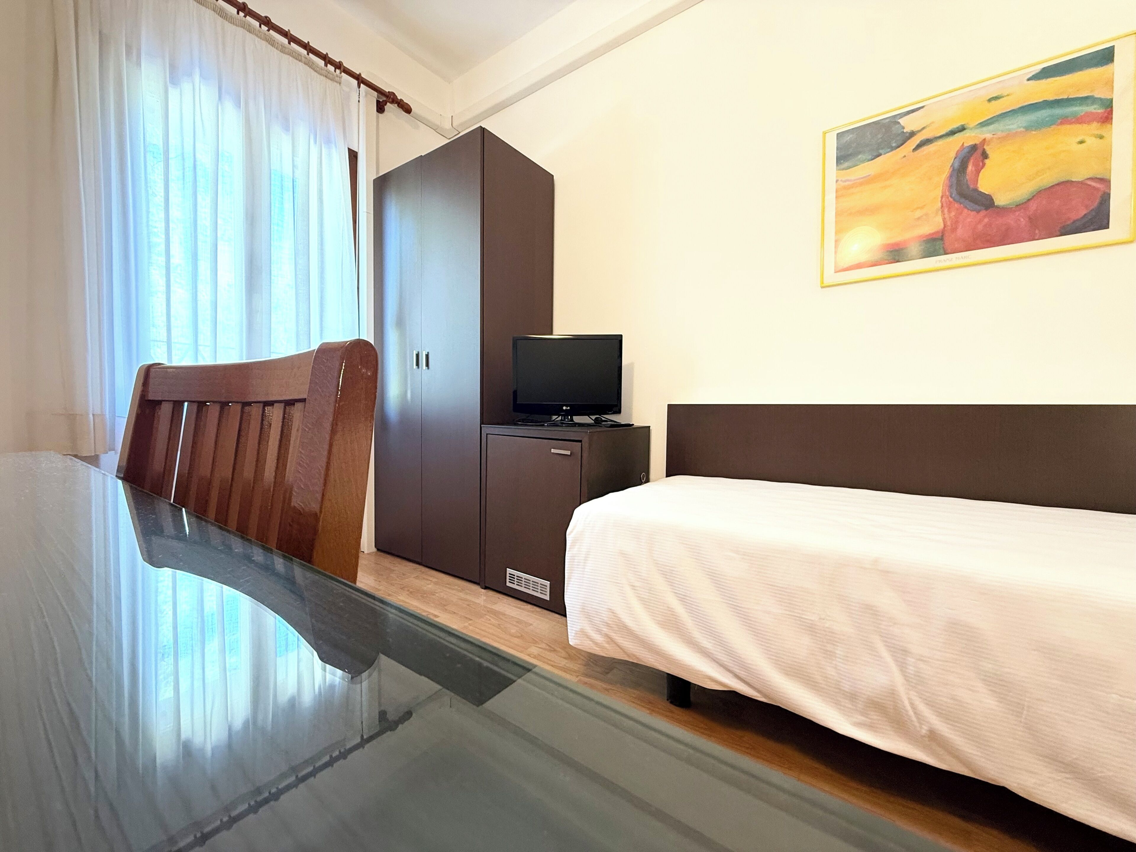 Habitación de hotel con cama, una silla de madera, televisor en un mueble y un cuadro en la pared.