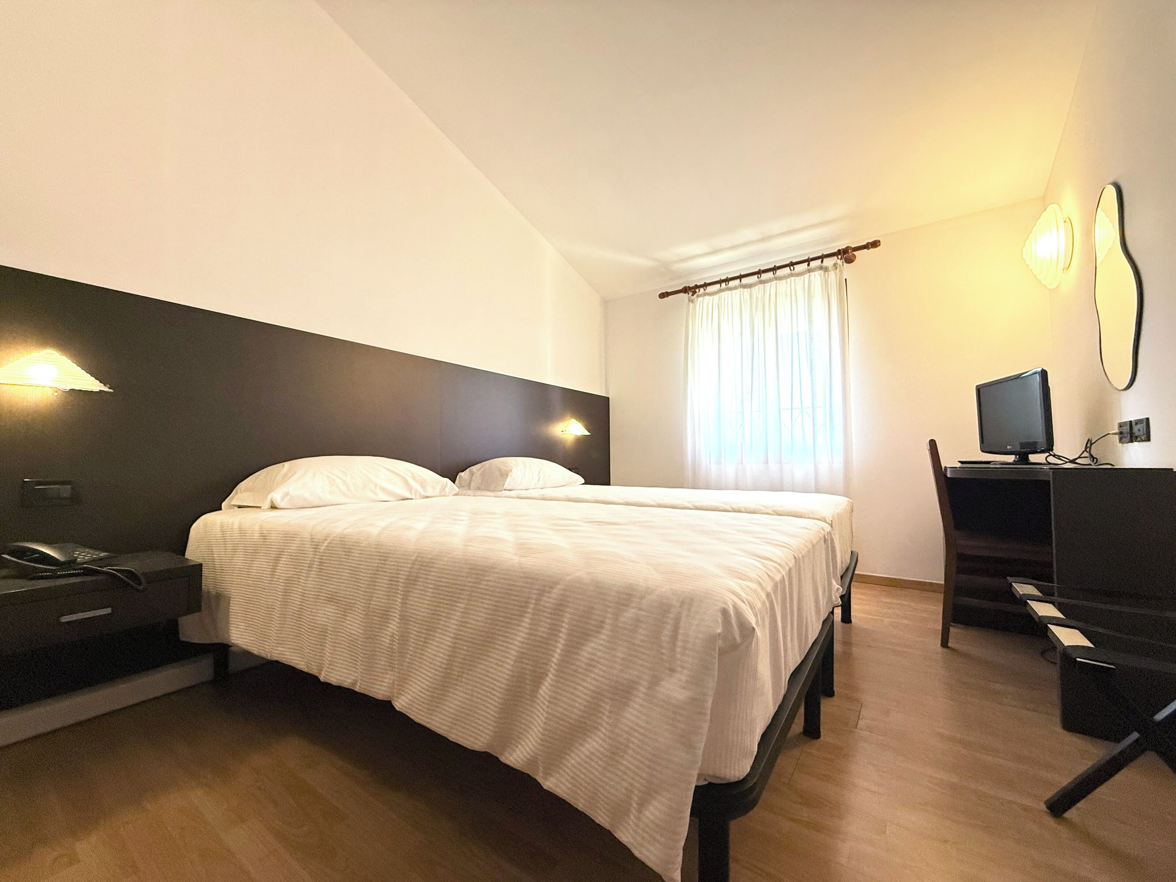 Habitación de hotel con cama, mesita de noche con teléfono, escritorio con computadora y ventana con cortinas.