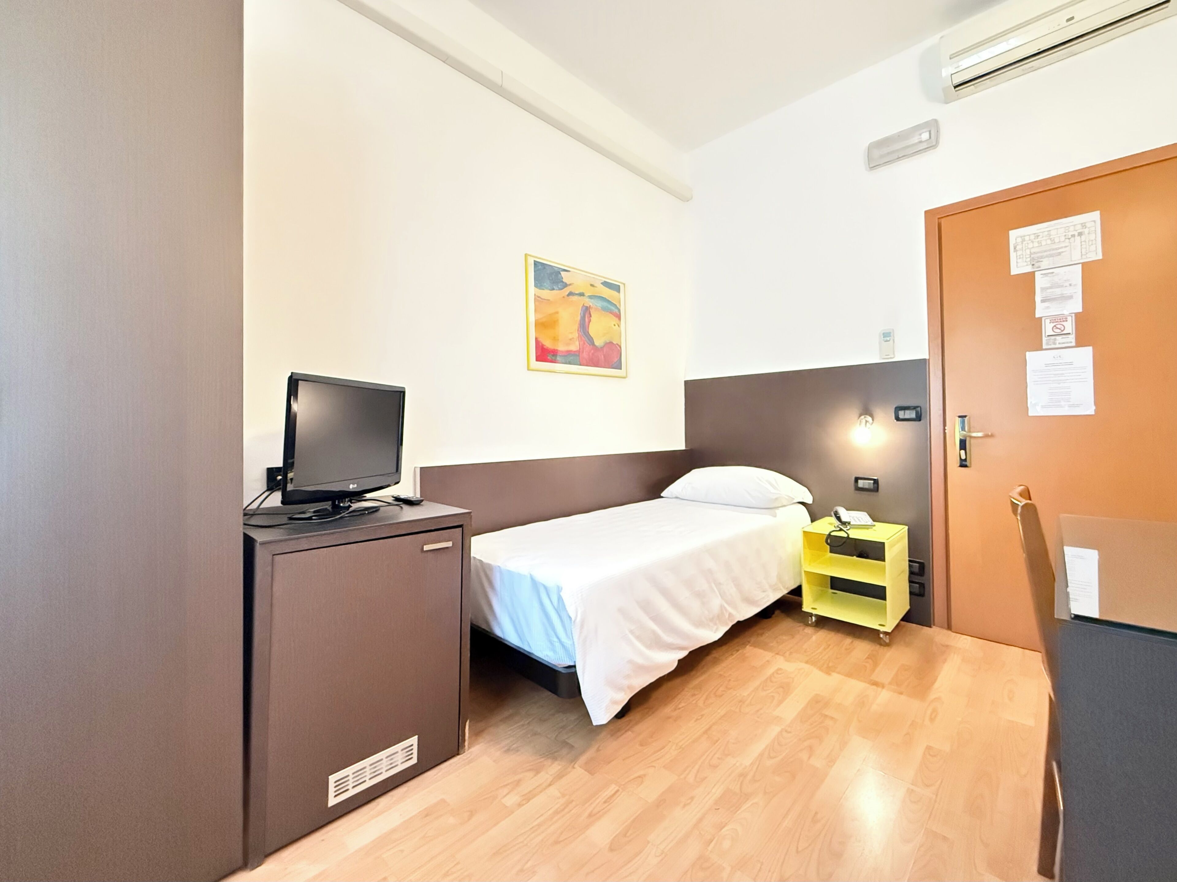 Una habitación de hotel pequeña con una cama, un escritorio con televisor, una mesita de noche con lámpara y una puerta con tablón de anuncios.