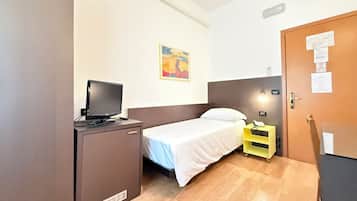 Una habitación de hotel pequeña con una cama, un escritorio con televisor, una mesita de noche con lámpara y una puerta con tablón de anuncios.