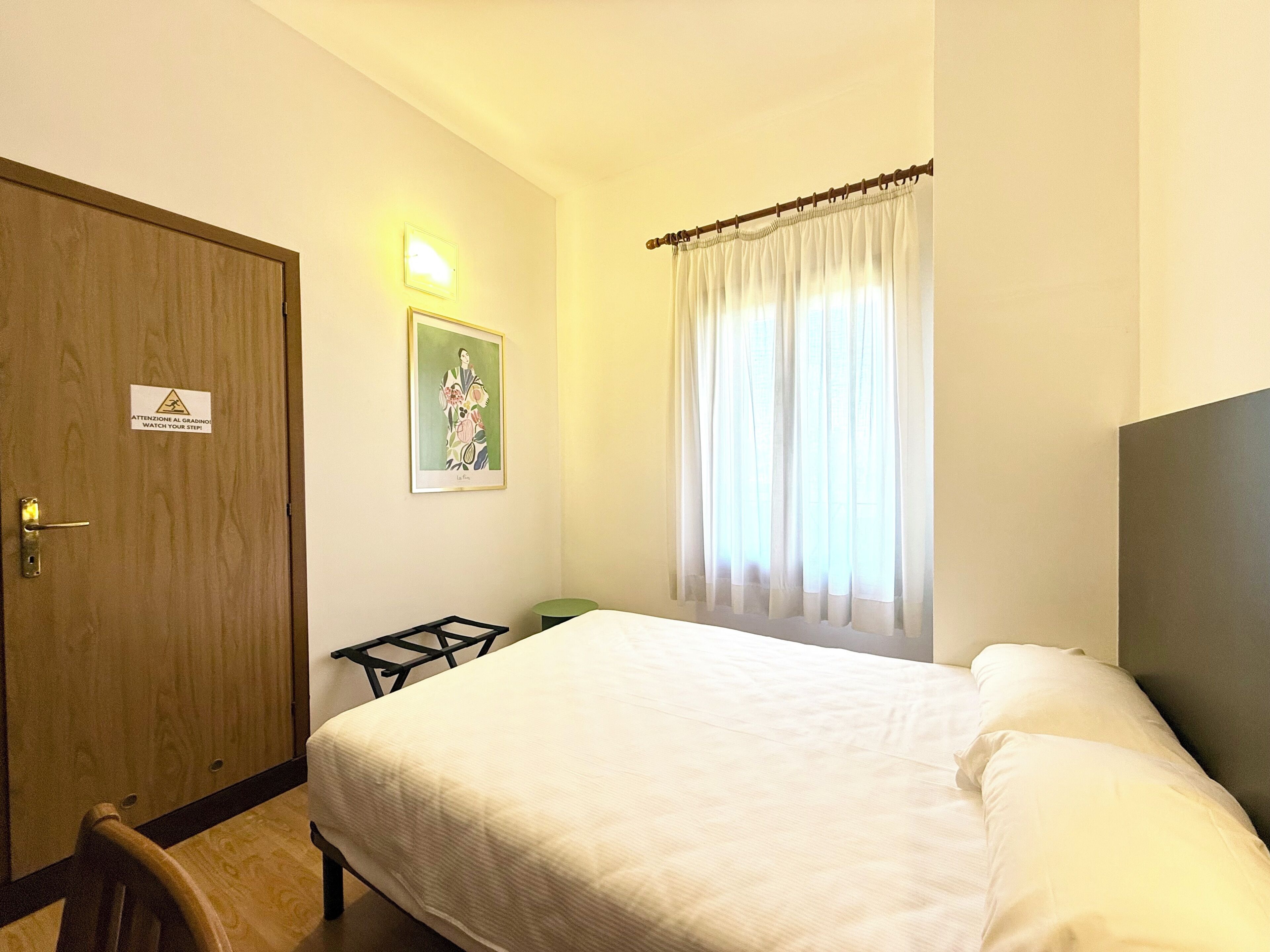 Une chambre d’hôtel avec un lit, une porte en bois, une petite table, un tableau encadré au mur et une fenêtre avec des rideaux.