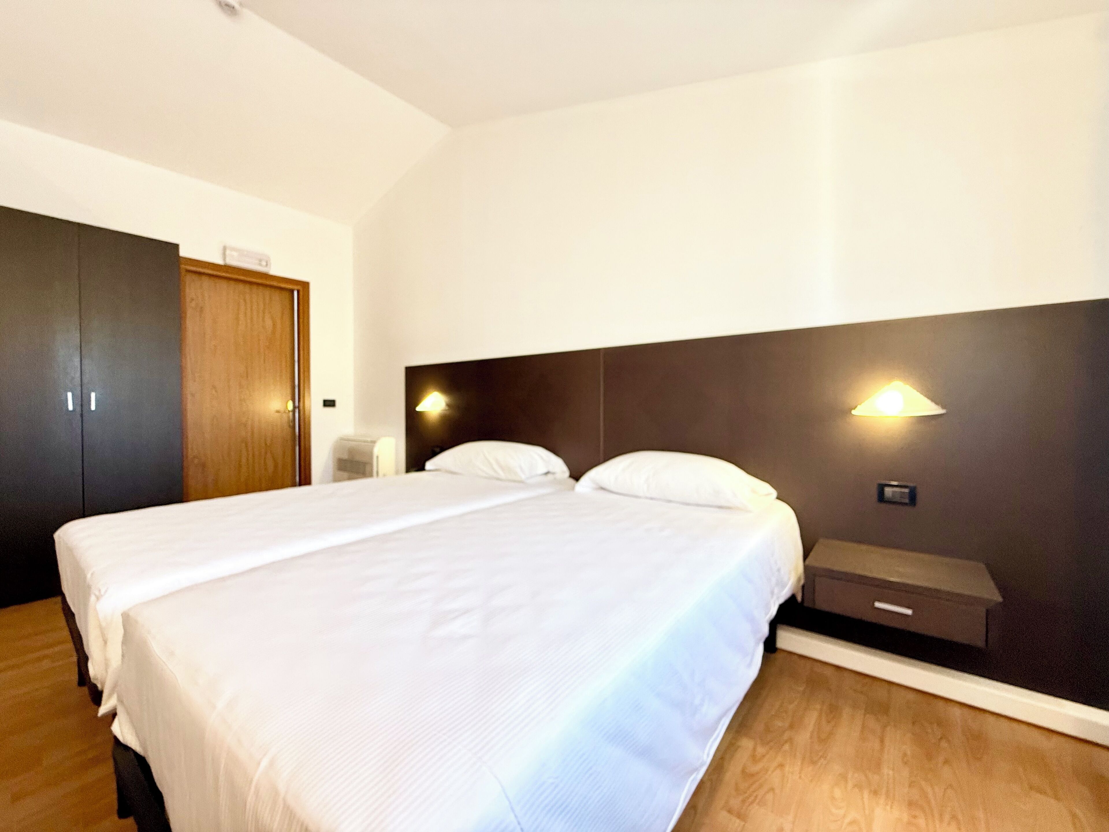 A hotel room with a large bed, a wooden wardrobe, and a nightstand with a lamp.