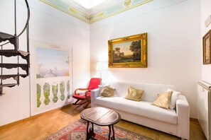 A living room with a white sofa, red rocking chair, and a small wooden table. A large framed painting hangs on the wall, and a radiator is visible on the right. - Vrbo Property (Roma)