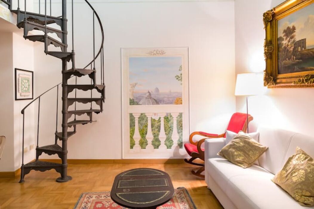 A room with a spiral staircase, a white sofa with decorative pillows, a red chair, a framed painting, and a door with a cityscape view.