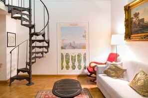 A room with a spiral staircase, a white sofa with decorative pillows, a red chair, a framed painting, and a door with a cityscape view. - Rome Apartment Trevi Spagna Charme, Barberini Metro Station (Roma)
