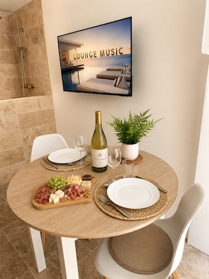 A dining area with a round table set for two, a bottle of wine, a potted plant, and a television displaying 'LOUNGE MUSIC' above it. - L'Escapade (Le Mans)