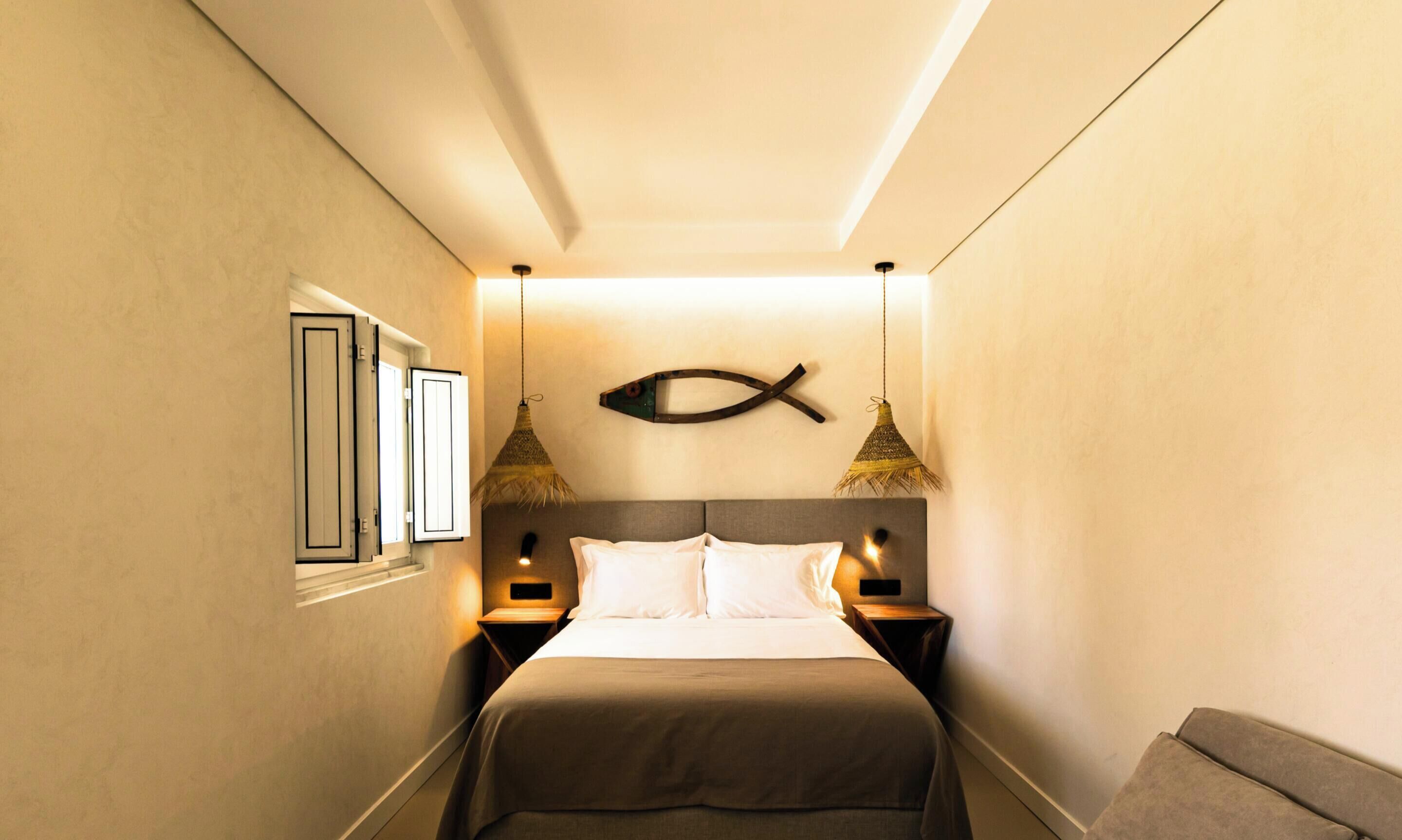 A narrow hotel room with a bed, bedside lamps, a wall-mounted fish decoration, and a window with blinds.