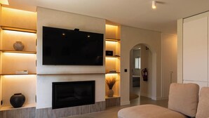 A modern living room with a flat-screen TV, fireplace, shelves with decorative items, and a sofa. - MyStay - Aunt Olinda (Porto Covo)