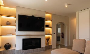 A modern living room with a flat-screen TV, fireplace, shelves with decorative items, and a sofa. - MyStay - Aunt Olinda (Porto Covo)