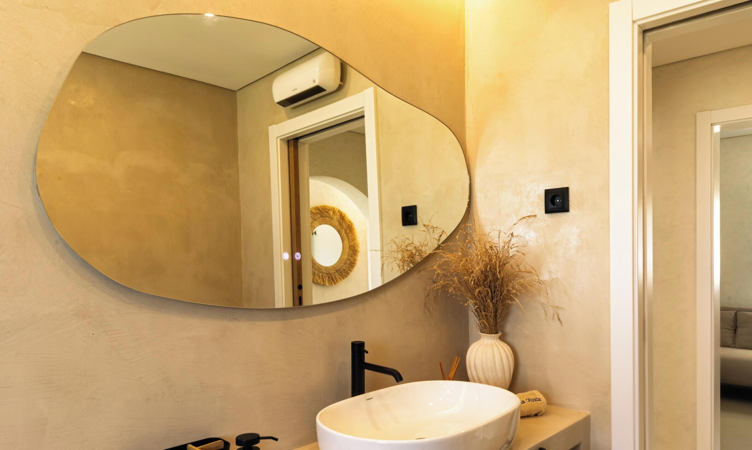 A bathroom with a large oval mirror, a white sink, and a wall-mounted air conditioning unit.