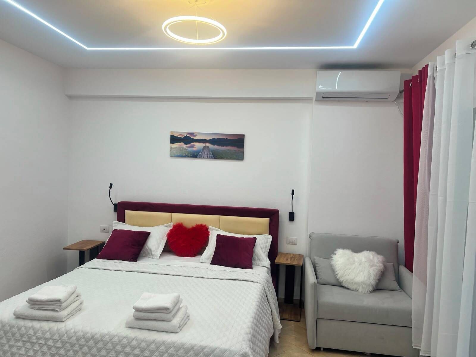 A modern hotel room with a bed, a bedside table, a chair, a painting on the wall, and an air conditioning unit.