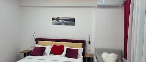 A modern hotel room with a bed, a bedside table, a chair, a painting on the wall, and an air conditioning unit.