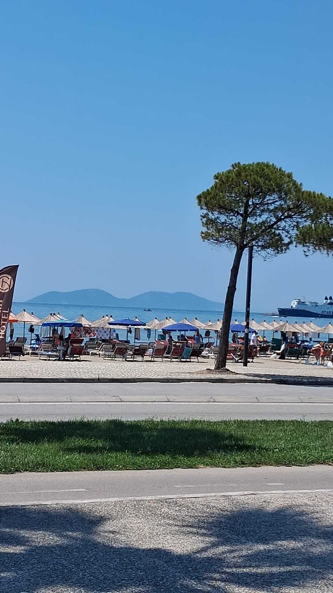 Un hôtel situé en bord de mer, doté d’une terrasse, de parasols et offrant une vue dégagée sur la mer et les montagnes en arrière-plan.