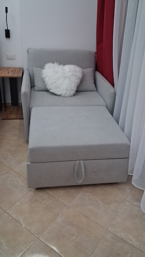 A grey sofa bed with a fluffy white pillow, a wooden side table, and a red curtain. - AA Holiday Apartment Longmore Vlore (Lungomare)