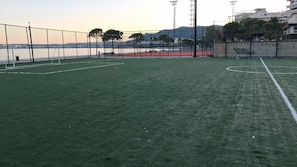 A sports field with a goalpost, fencing, and a view of a body of water and buildings in the background. - AA Holiday Apartment Longmore Vlore (Lungomare)