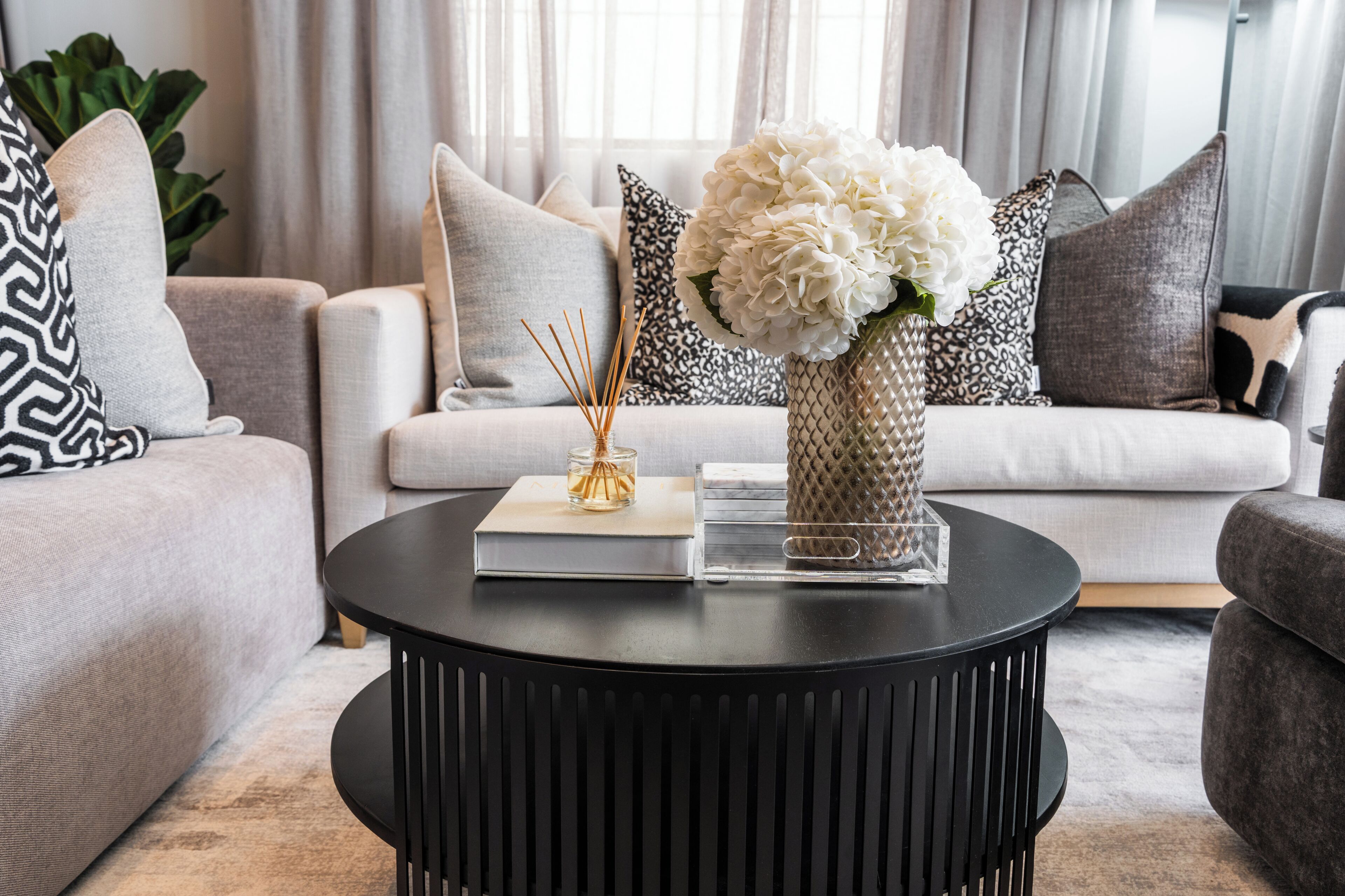 En moderne stue med en sofa, et rundt sort sofabord og dekorative genstande, herunder en vase med blomster og en bog.