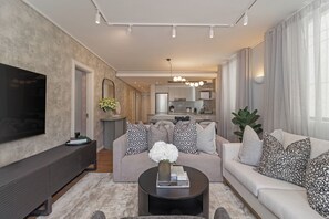 Ein modernes Wohnzimmer mit Flachbildfernseher, Couchtisch mit Dekoration, Sofa mit gemusterten Kissen und einer Küche im Hintergrund.