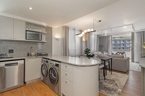 Eine moderne Küche mit integrierten Geräten, ein Essbereich mit grauem Sofa und Blick auf ein Gebäude durch das Fenster.