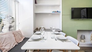Un comedor con una mesa blanca dispuesta para cuatro personas, copas, platos y cubiertos. Hay estanterías encima de la mesa y un televisor montado en la pared.