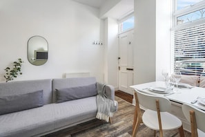 A modern living room with a grey sofa, a dining table set for two, and a large window with blinds. - Modern - 2BR Flat near Easter Road Stadium (Edinburgh)