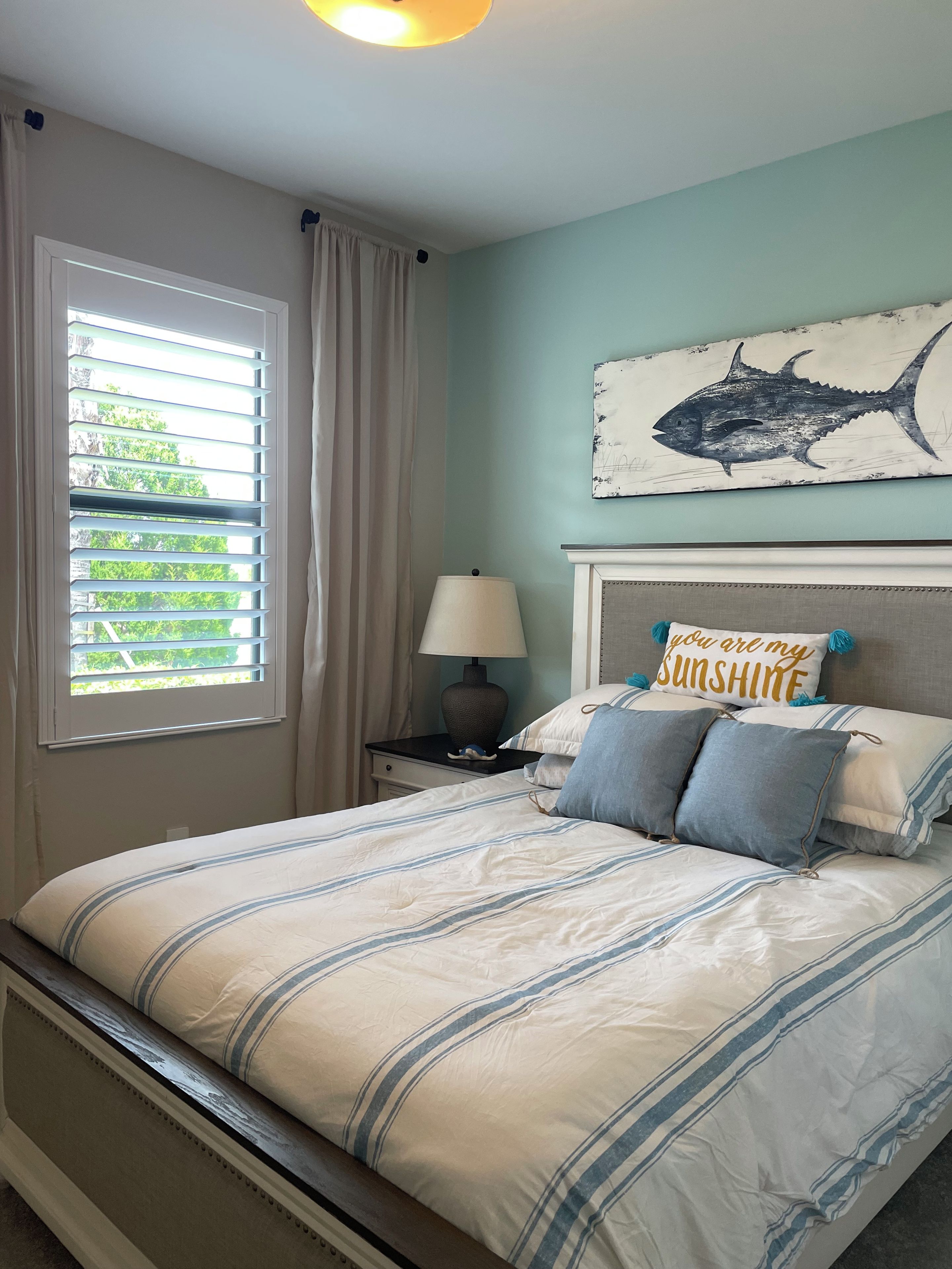 Une chambre à coucher comprenant un lit, une table de chevet, une lampe, une fenêtre avec des rideaux et une œuvre d’art représentant des poissons accrochée au mur.