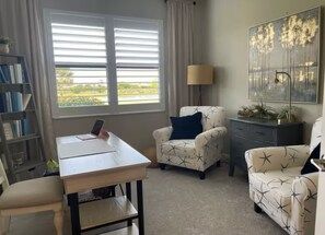 A room with a desk, two armchairs, a lamp, a bookshelf, and a large window with blinds. - Charming 2-bedroom Villa on water w/ Many Amenities in gated Lakewood Ranch (Bradenton)