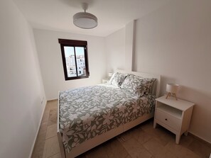 Ein Schlafzimmer mit einem Bett, einem Nachttisch, einer Lampe, einem Fenster und einem Deckenventilator.