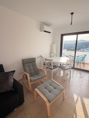 A modern living room with a sofa, armchair, ottoman, and dining set. An air conditioning unit is mounted on the wall, and a large window provides a view of the outdoors. - 3 bedroom, central apartment with communal pool and stunning views (Frigiliana)