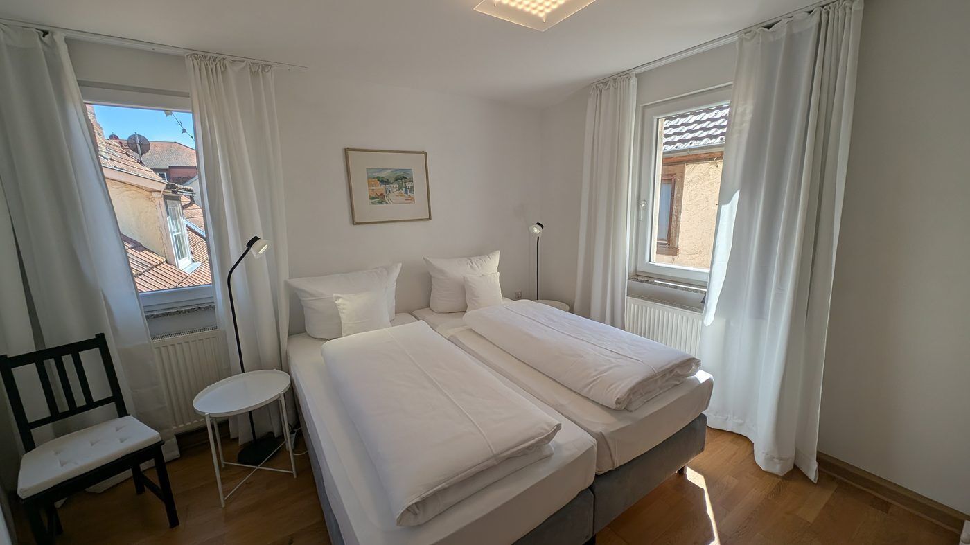 Un dormitorio con cama, una silla, una mesita y dos ventanas con cortinas.