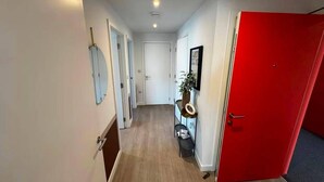 A hallway with a red door, a white door, and a door with a circular mirror. There is a small table with a plant and a framed picture. - Cozy London Apartment With 2 Bedrooms (London)