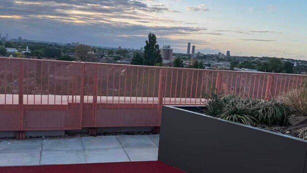 A rooftop terrace with a red carpet, a metal fence, and a view of a cityscape with buildings and trees. - Cozy London Apartment With 2 Bedrooms (London)