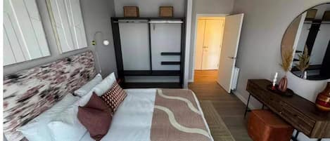 Un dormitorio con cama, mesita de noche, cómoda, espejo, armario y una puerta que da a otra habitación.
