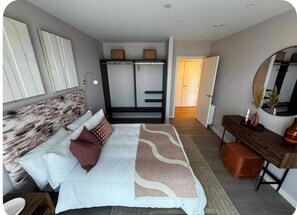 Ein Schlafzimmer mit Bett, Nachttisch, Schminktisch, Spiegel, Kleiderschrank und einer Tür zu einem weiteren Raum.