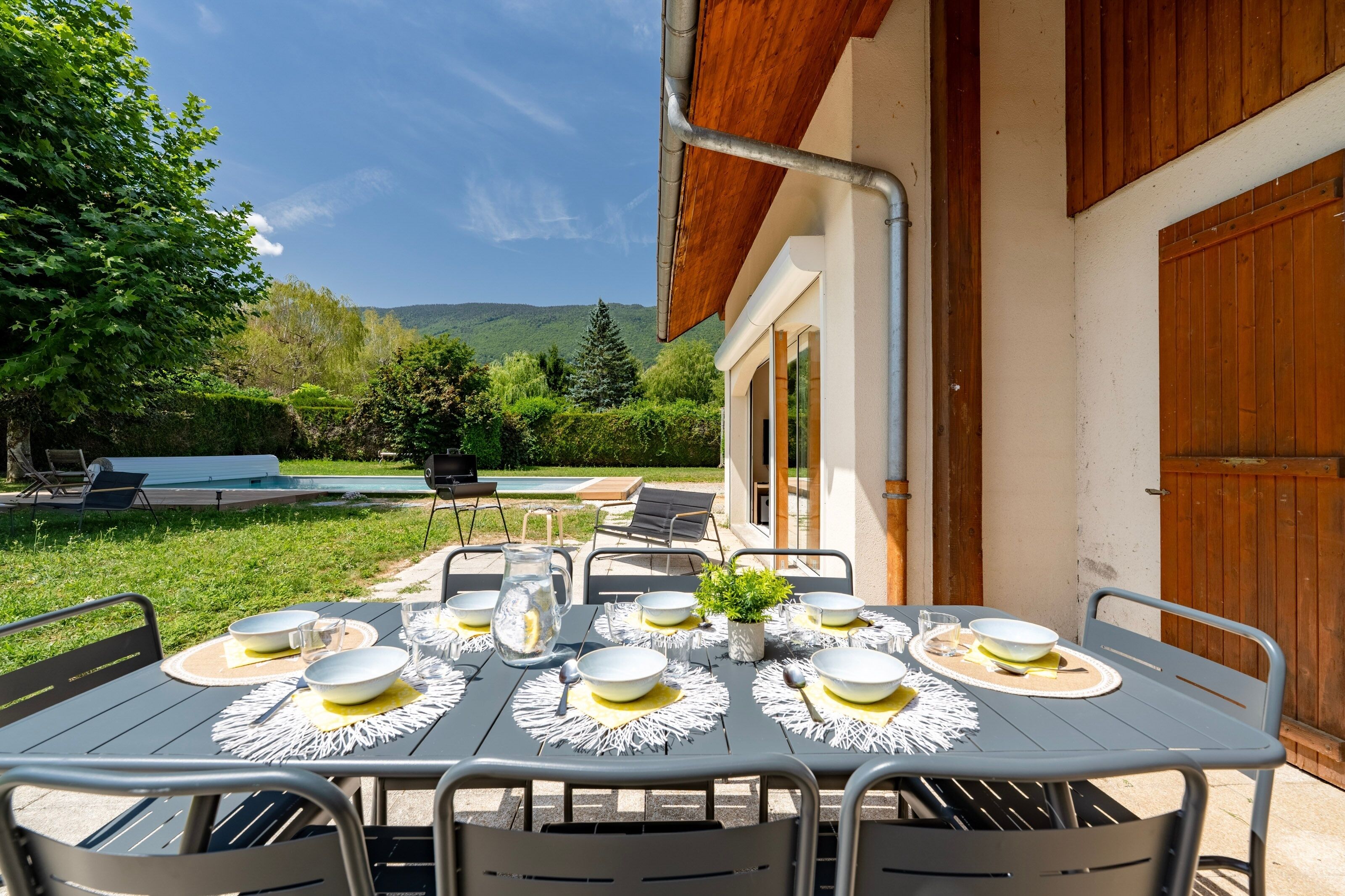 Un espace extérieur aménagé pour les repas, avec une table dressée pour quatre personnes, entourée de chaises, et offrant une vue sur une piscine et une végétation luxuriante en arrière-plan.