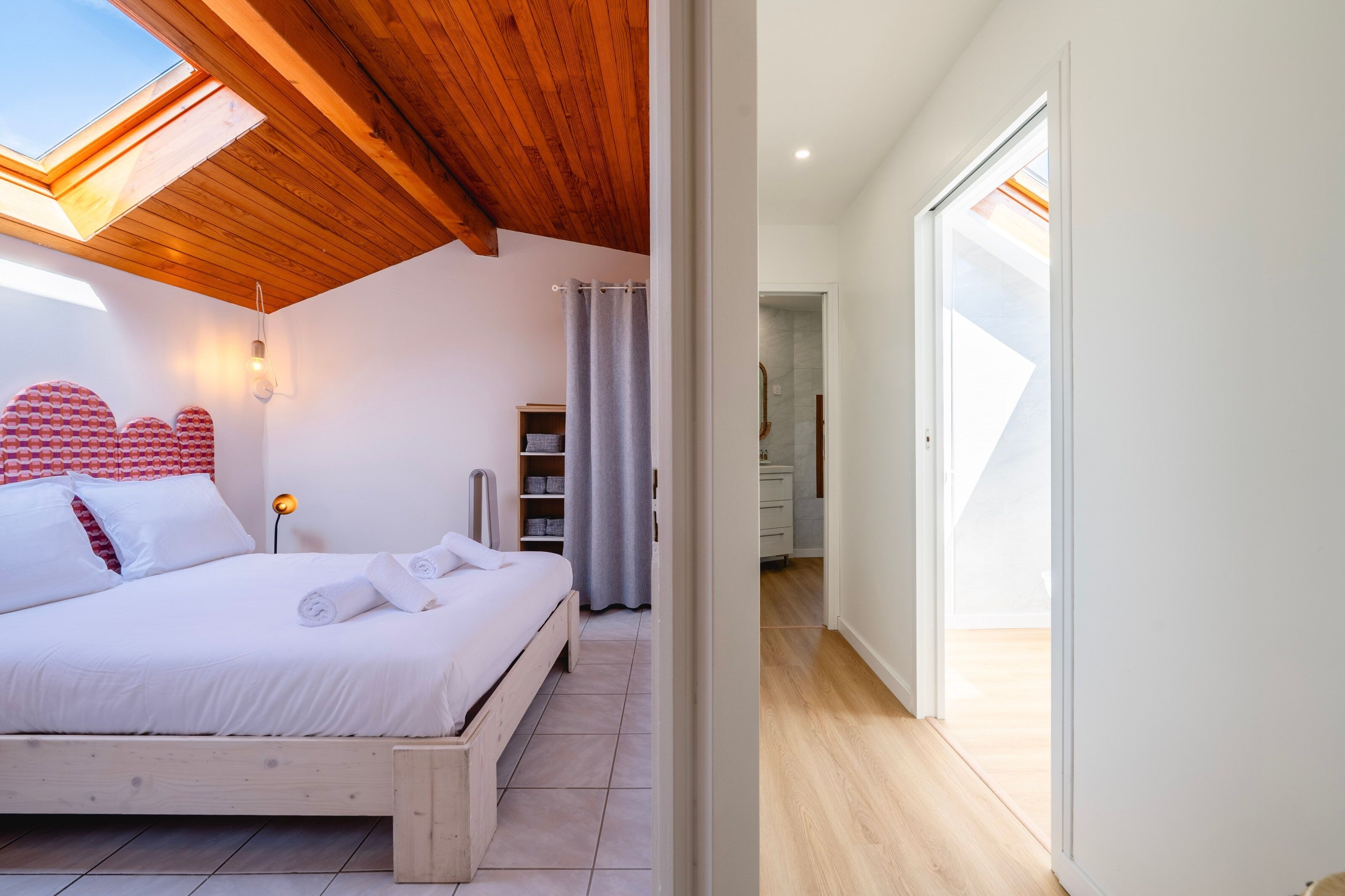 Une chambre avec un puits de lumière, un lit recouvert de draps blancs, une tête de lit en bois et une table de chevet avec une lampe.