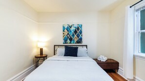 Un dormitorio con cama, una mesita de noche con lámpara, una pequeña mesa con toallas, un cuadro en la pared y una ventana con cortinas.