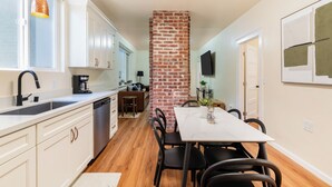 Una cocina moderna con armarios blancos, una pared de ladrillo decorativa, una zona de comedor con mesa blanca y un televisor montado en la pared.