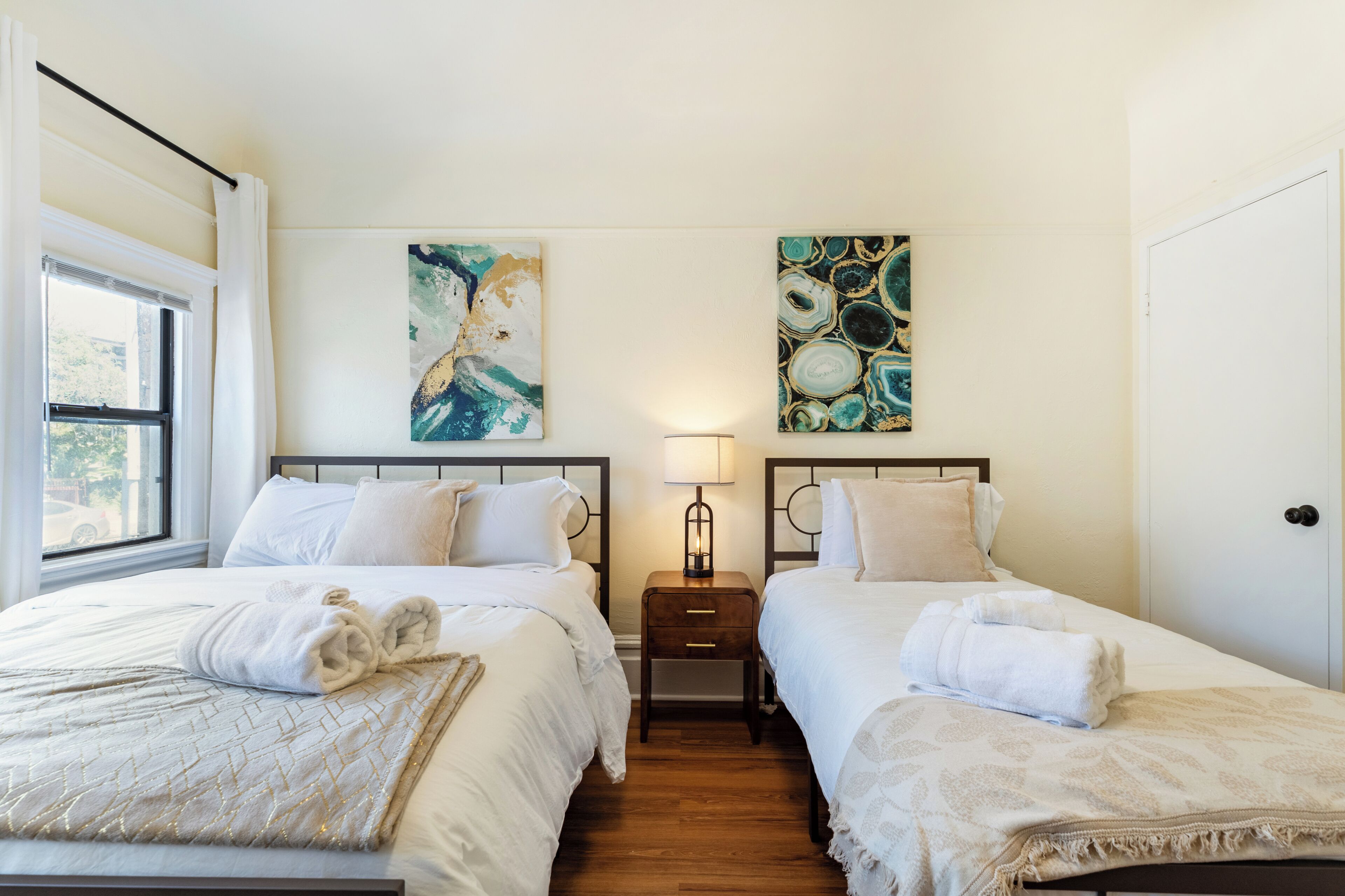 Une chambre d’hôtel avec deux lits, une table de chevet avec une lampe, des œuvres d’art aux murs et une fenêtre avec des rideaux.