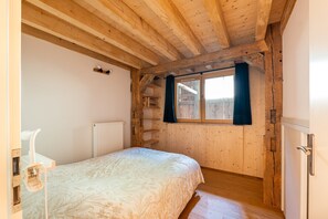 A bedroom with a wooden ceiling, a bed, a window with curtains, a radiator, and a white chair. - Le Refuge de La Forge - Charmante ferme rénovée (La Forge)