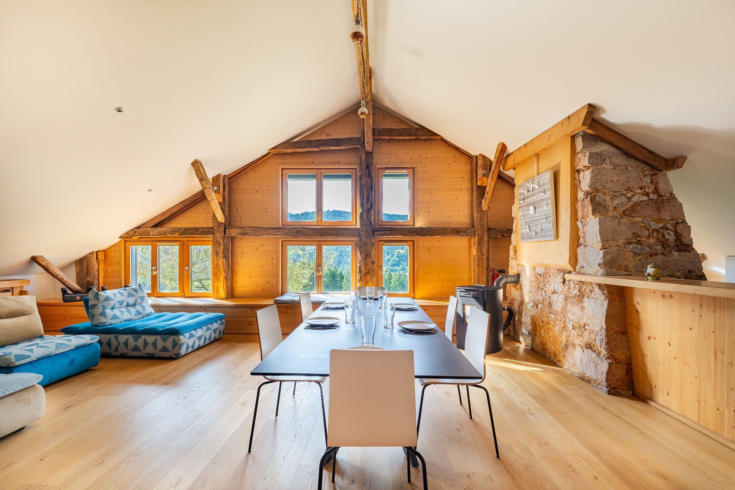 A spacious loft-style room with a high ceiling, a long dining table set for four, and a stone fireplace.