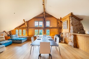 A spacious loft-style room with a high ceiling, a long dining table set for four, and a stone fireplace. - Le Refuge de La Forge - Charmante ferme rénovée (La Forge)