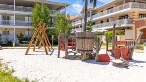 A beachfront hotel with a sandy beach area, wooden play structures, and a multi-story building with balconies. - Instant Evasion - Piscine Partagée (La Teste-de-Buch)